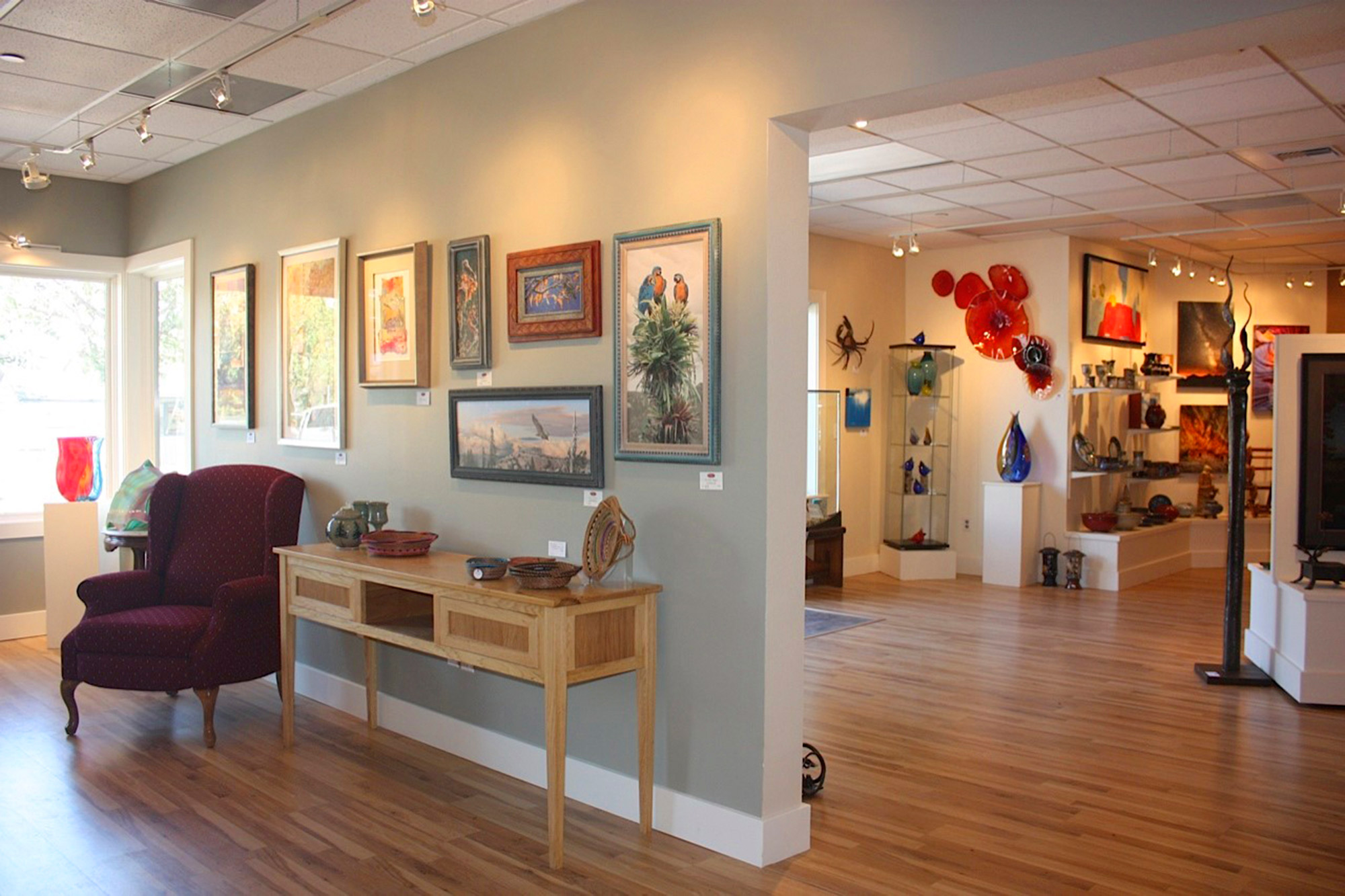 Gallery room with wooden floors, artwork on walls, and a maroon chair.