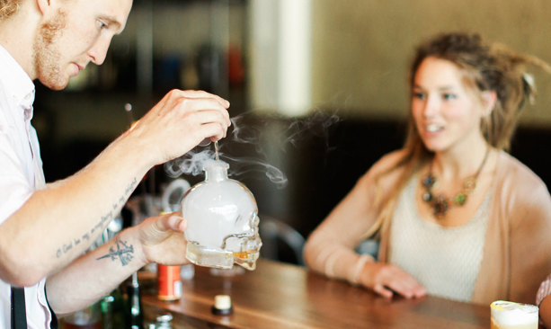 bartender crafting a cocktail with smoke while seated patron looks on