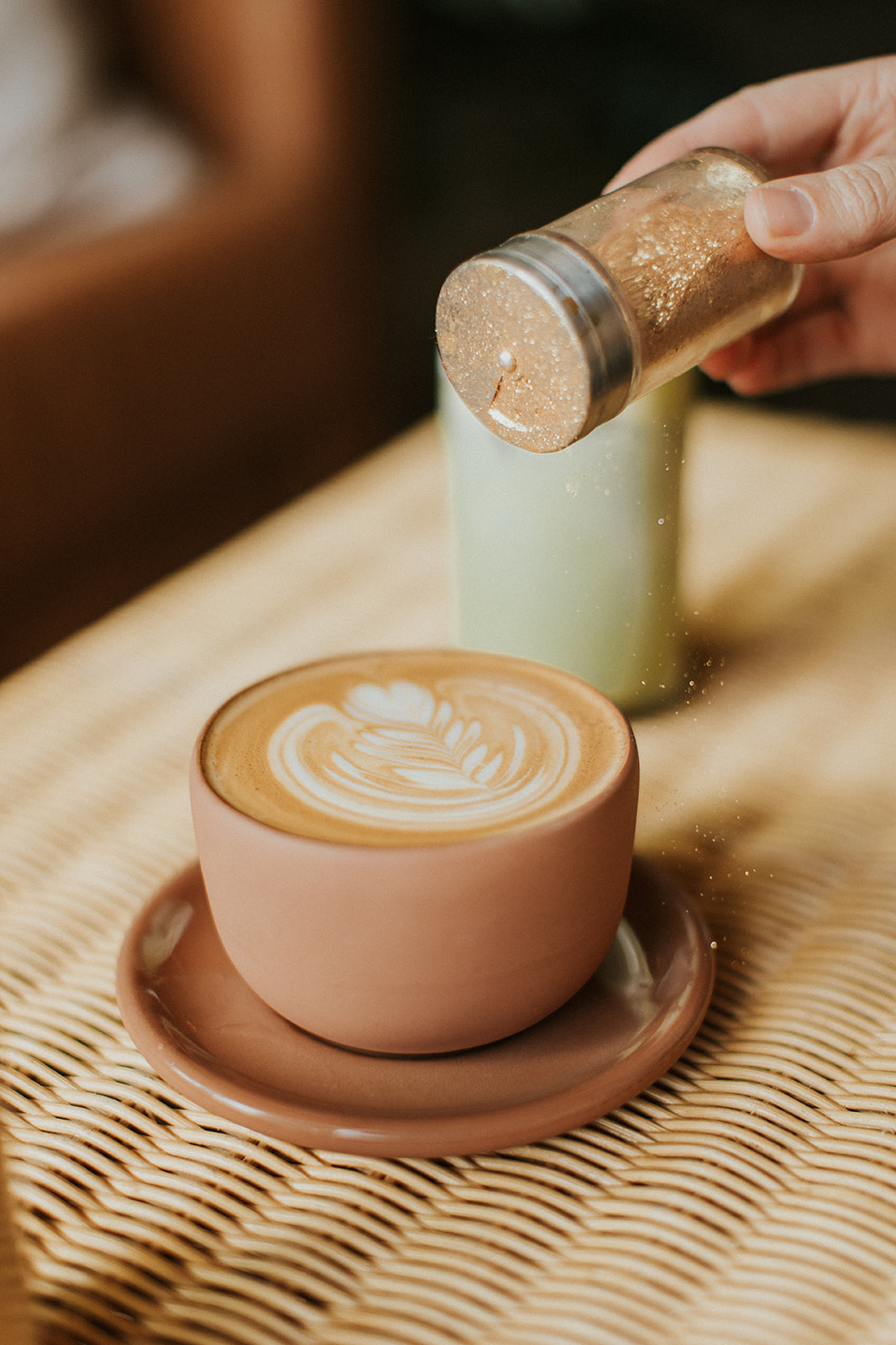 Hand holding shaker of edible rose gold glitter and pouring it on an espresso in a terra cotta cup on a wicker table at Golden Coffee In Prineville, Oregon