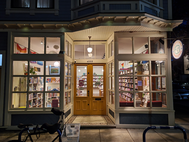 A  warmly lit bookstore storefront at night, featuring large glass windows displaying shelves filled with books and colorful decorations.