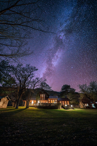 Nighttime rural scene featuring a house with illuminated windows under a clear sky filled with stars.