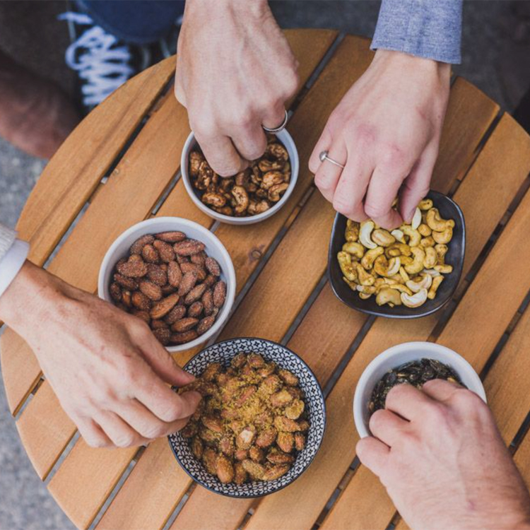 five bowls of nuts on a table with four hands reaching for them