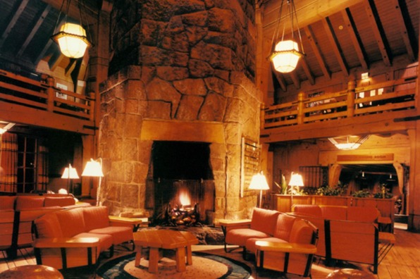 A rustic lodge interior featuring a large stone fireplace as central focal point surrounded by orange cushioned wooden chairs and small tables.