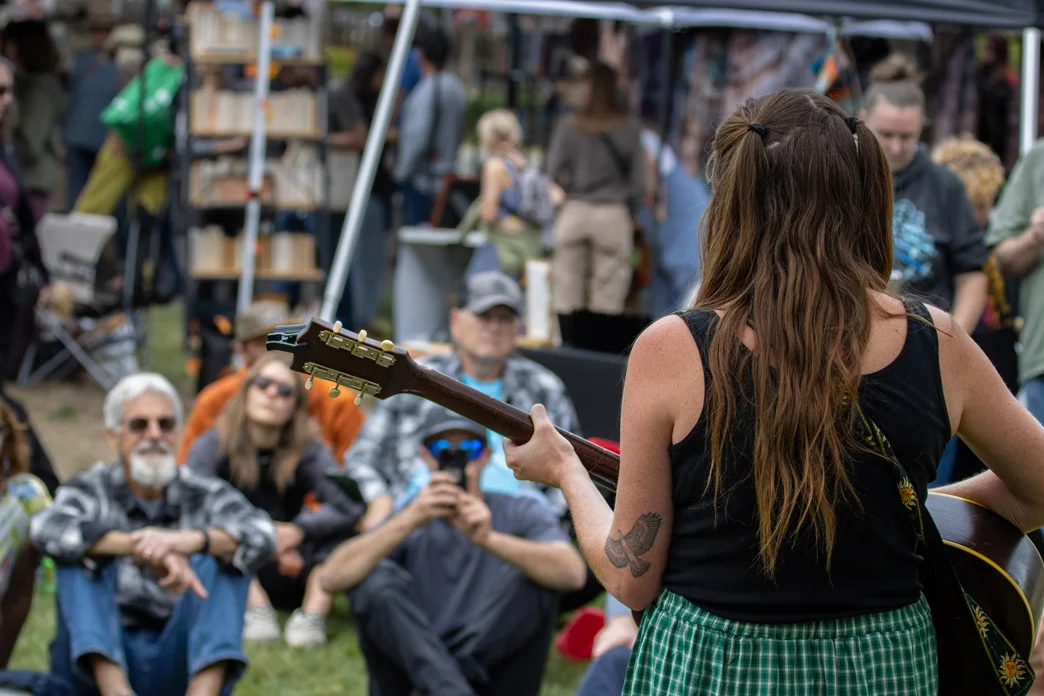 Seated crowd watches woman performing with acoustic guitar at outdoor festival.