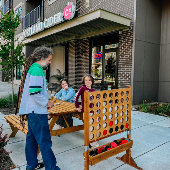 family plays large Connect4 game on sidewalk with Portland Cider Company building in background