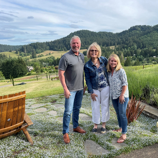three people smile with vineyard behind them