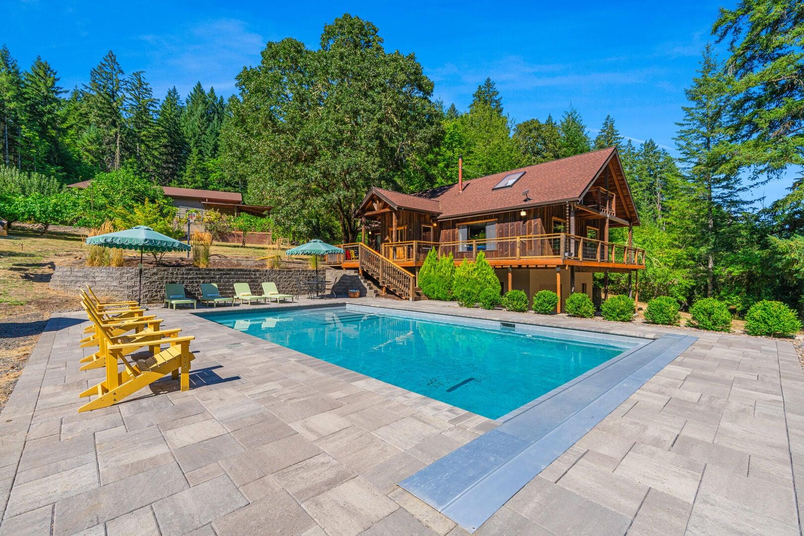 An outdoor swimming pool area adjacent to a wooden cabin surrounded by tall trees and greenery.