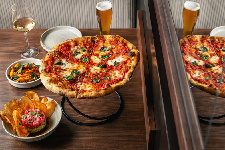 A dining setup featuring a Margherita pizza on a black metal stand, accompanied by a glass of white wine, a glass of beer, and two small bowls of appetizers on a wooden table.