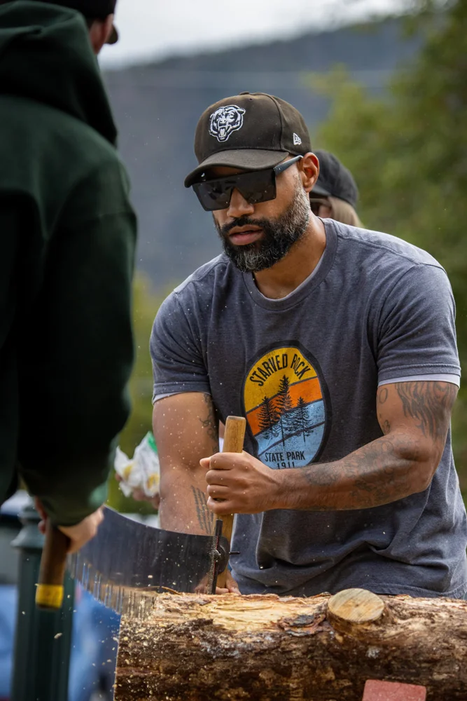 Man participates in a sawing race at an outdoor festival.