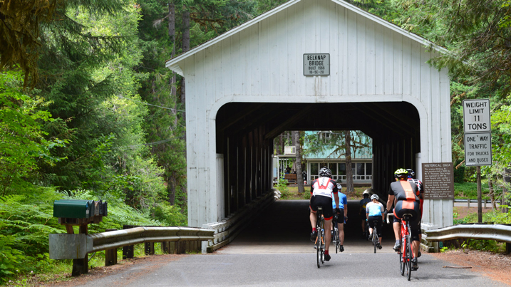 Group of bicyclists ride toward a covered bridge.