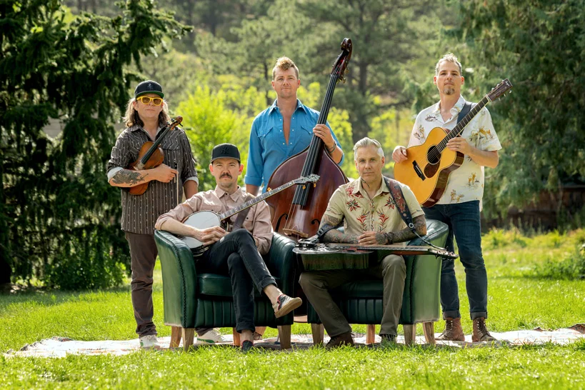 Five members of a band pose with their instruments and a couch in a wooded outdoor setting.