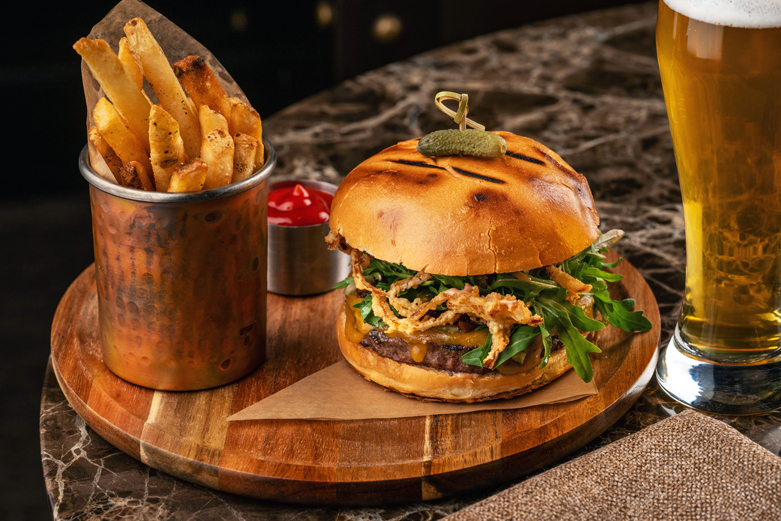 Gourmet burger meal featuring a toasted bun topped with a small pickle, crispy fried onions, and fresh greens, served on a wooden board next to a glass of beer.