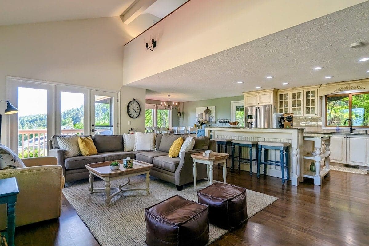 An open-concept living room and kitchen with sectional, yellow/white pillows, wood coffee table, and two leather ottomans.