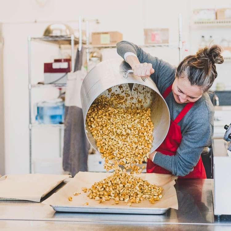 person in commercial kitchen transferring flavored nuts from large pot to sheet tray