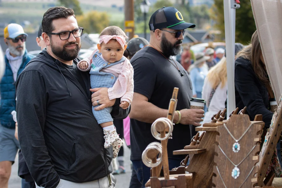 Father holds baby daughter at outdoor festival vendor tent.