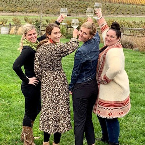 four people each raising a wine glass with bodies facing away from camera but heads turned to smile at camera