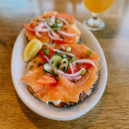 bagel and lox on an oval plate next to a stemmed glass of cider