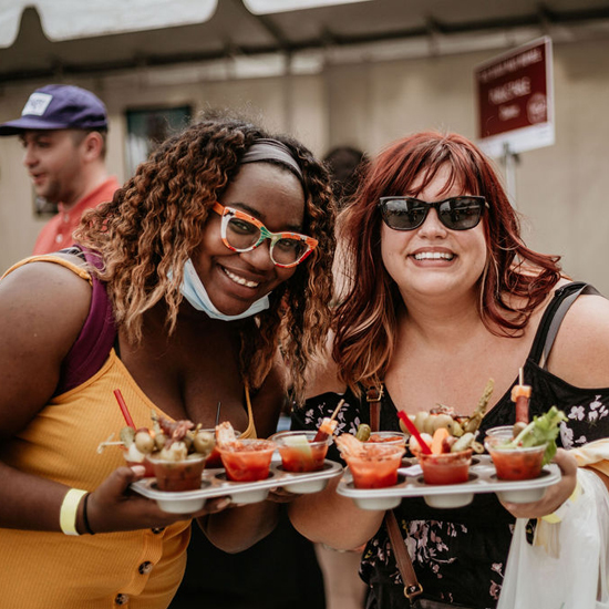 two people smile while each holding a taster tray of six bloody mary beverages