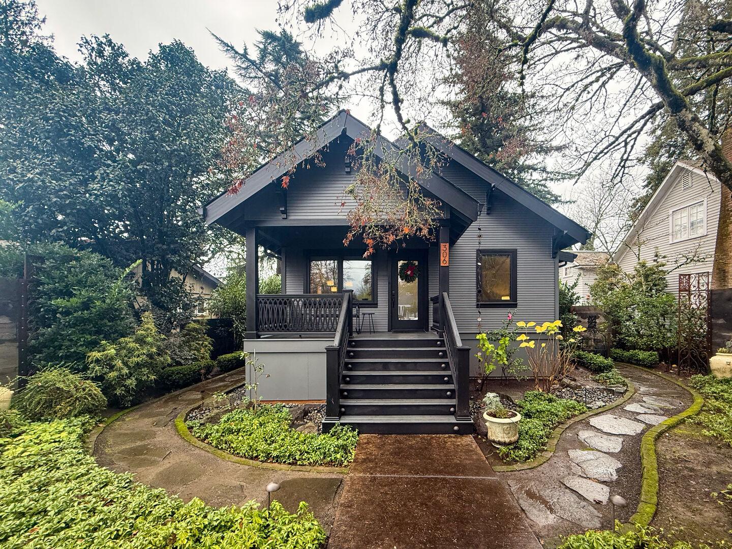 Gray bungalow-style house with a front porch and black stairs, surrounded by lush greenery and a curved stone pathway.