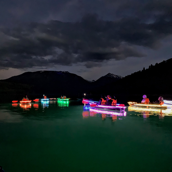 group of kayakers paddle on a lake under the night sky in illuminated kayaks