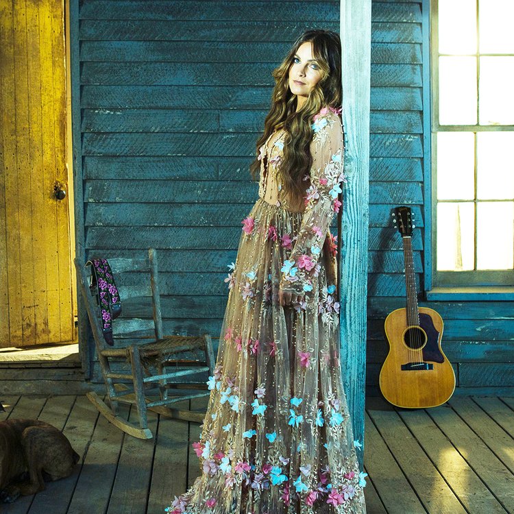 woman wearing long dress leans against porch pillar will acoustic guitar and rocking chair in background