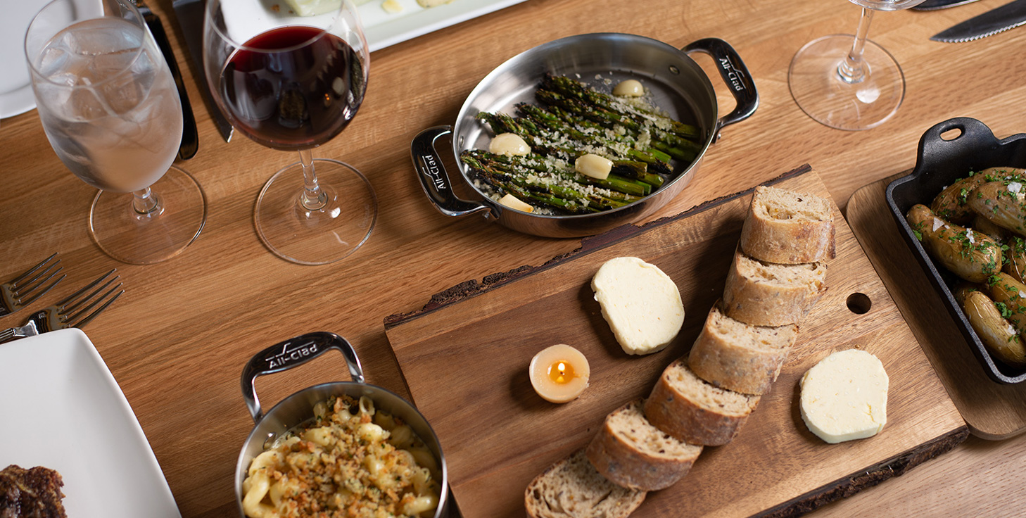 dinner dishes presented in cast iron pans and a bread plate on a wooden table top with glasses of wine and water