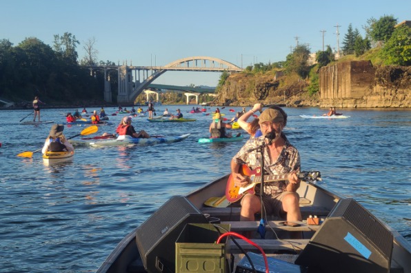 Person in a boat plays a guitar and sings into a microphone while a group of people kayak in the background.