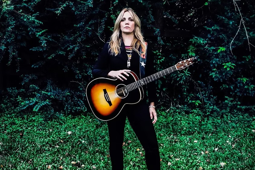 Woman holding acoustic guitar in front of lush greenery.