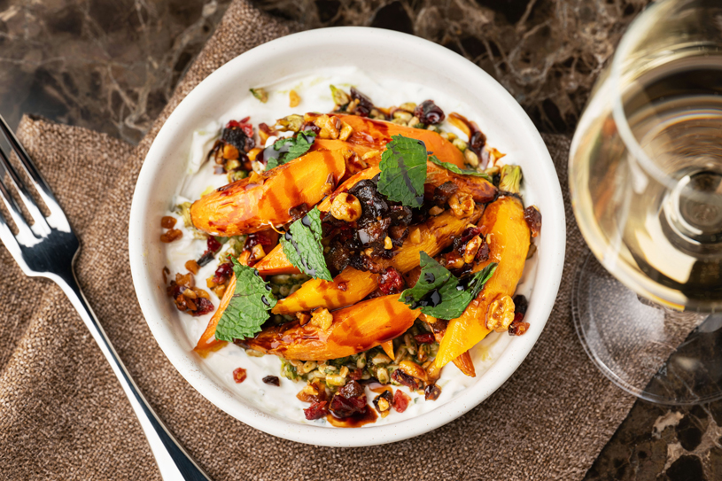 A white bowl containing roasted carrots, garnished with fresh mint leaves and a crunchy topping, served on a brown cloth napkin with a fork and a glass of white wine nearby.
