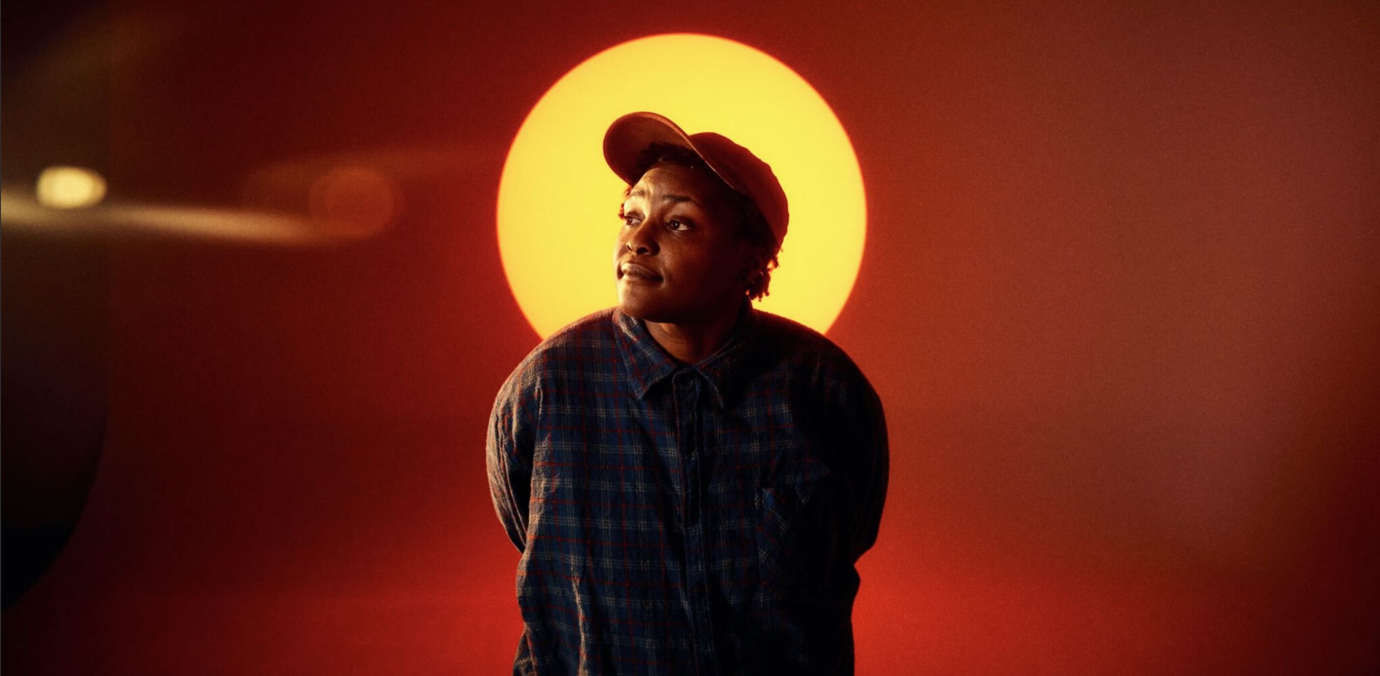 Artist Joy Oladokun wears a cap and flannel standing in front of yellow lighted circle
