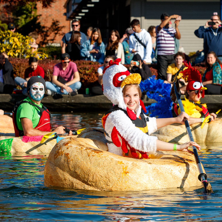 three people in costumes paddle in hollowed out giant pumpkins