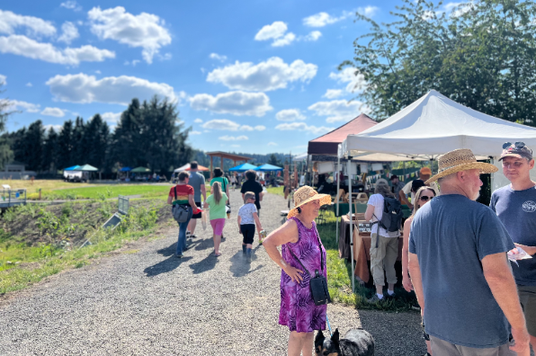 An outdoor market on a sunny day with blue sky and scattered clouds, showing people walking along a gravel path and browsing vendor tents.