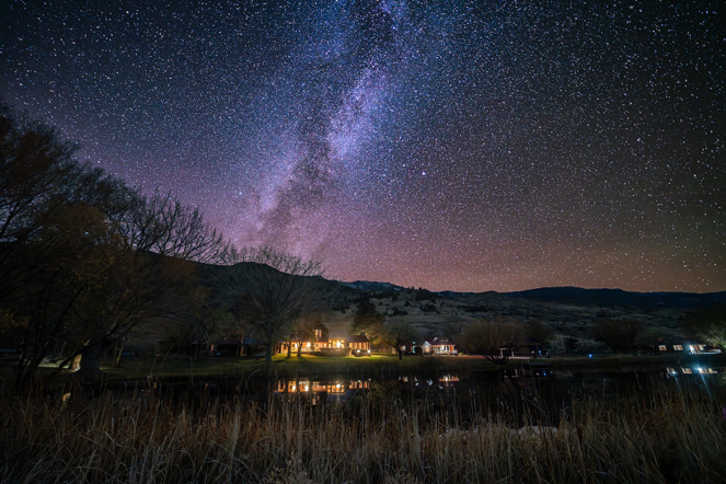 Starry night sky above a rural landscape with illuminated houses reflecting on a calm body of water.