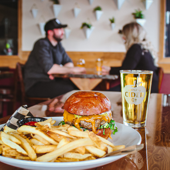 plate of cheeseburger and fries with pint of cider on table with couple seated at table in background