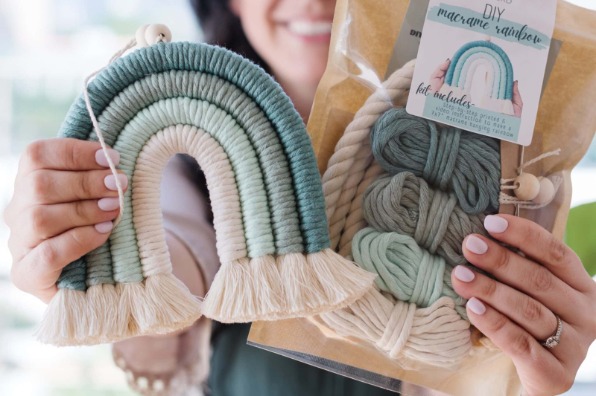 A person holding a completed macrame rainbow craft and a DIY macrame rainbow kit with yarn in shades of green, beige, and blue.