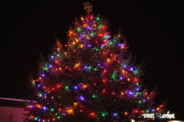 A large Christmas tree decorated with multicolored string lights in red, green, blue, yellow, and purple, topped with a glittery snowflake.