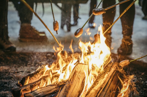 Several people roasting sausages on sticks over an open campfire with visible flames and burning logs.