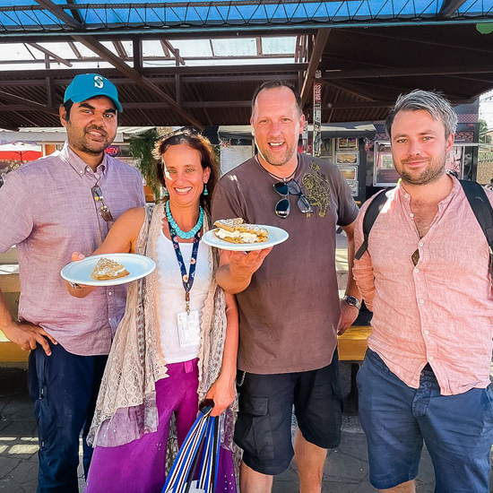 four people smile while at an outdoor food cart pod with two in center each holding up a plate with a cream filled dessert