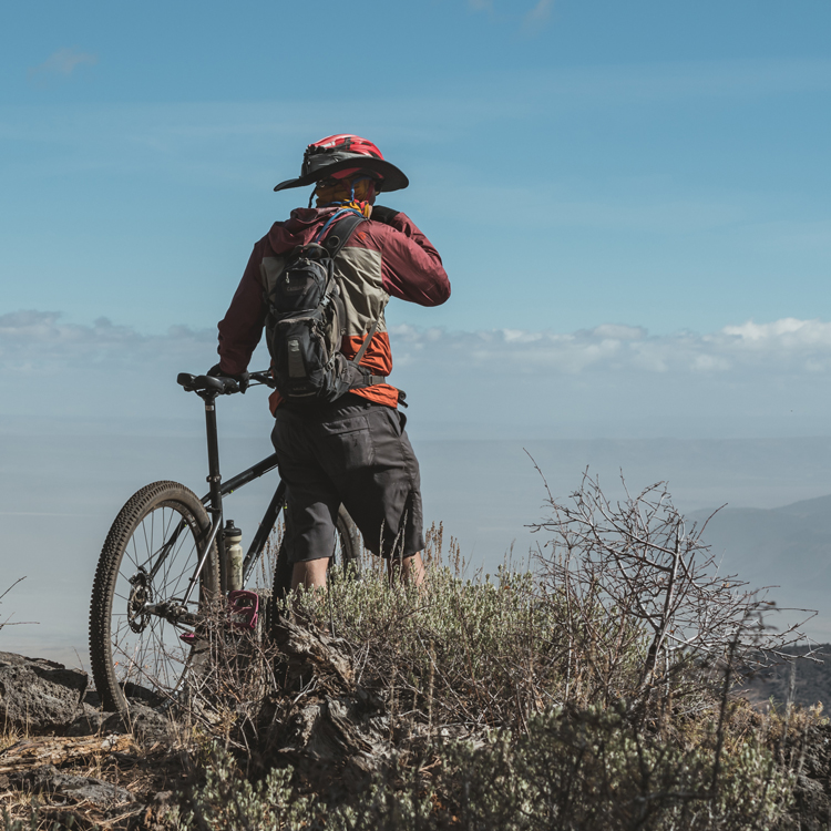 bicyclists wearing hat facing away from camera stands next to mountain bike while taking in a scenic view
