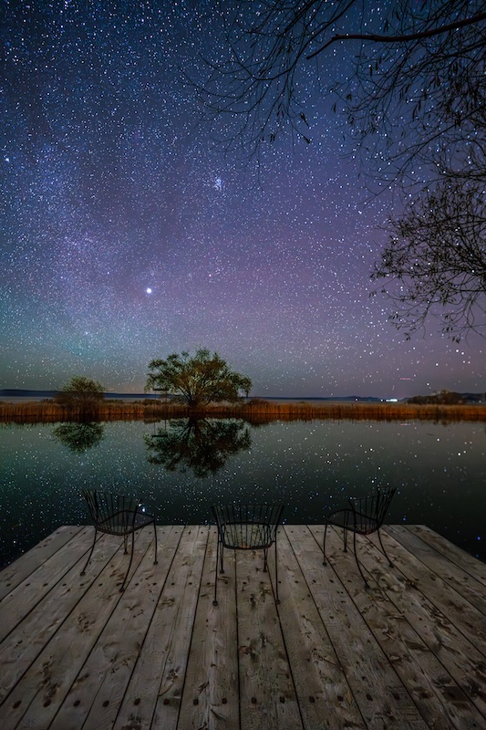 A wooden dock with three empty chairs overlooking a calm lake under a star-filled night sky.