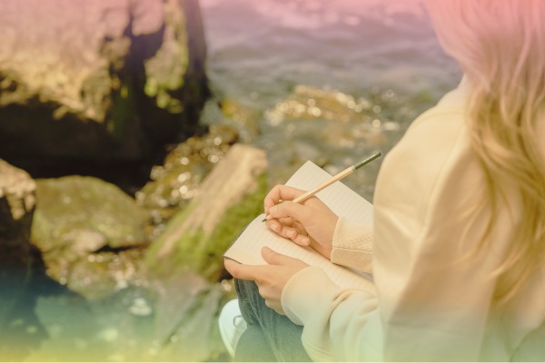 Woman writes in a journal while seated next to water.