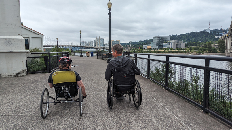 a person using a wheelchair and a person riding an adaptive bike on a riverfront path