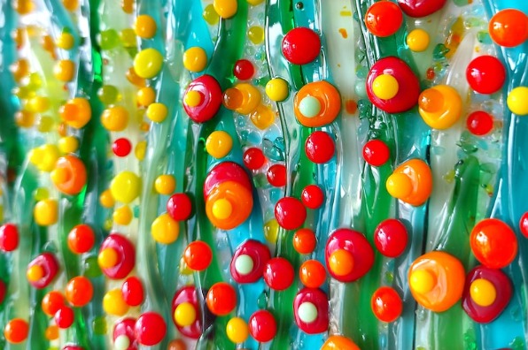 Close-up of colorful glass art featuring raised circular dots in red, orange, yellow, and white on a textured blue-green background.