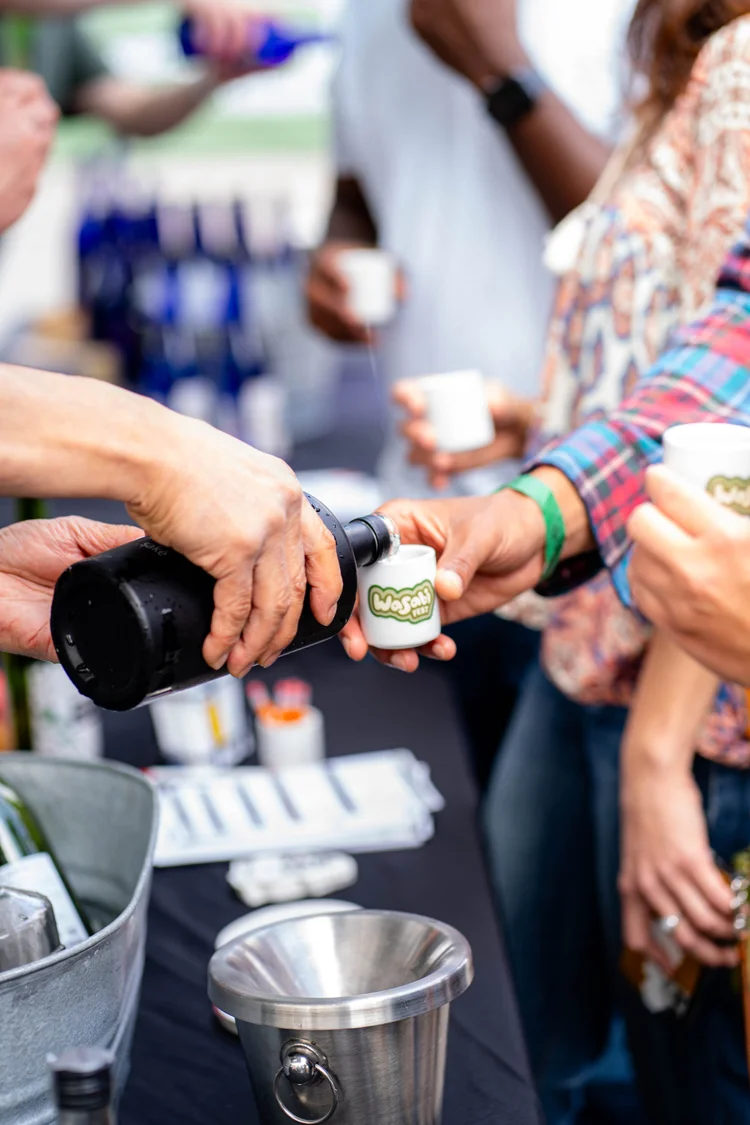 A person pours a saké from a bottle into a small white tasting cup labeled with WasabiFest logo.