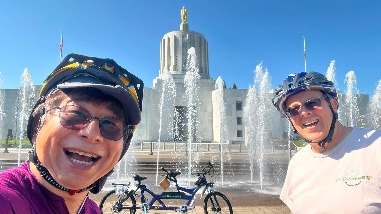 Two people wearing bike helmets smile in front of a fountain at Oregon State Capitol.