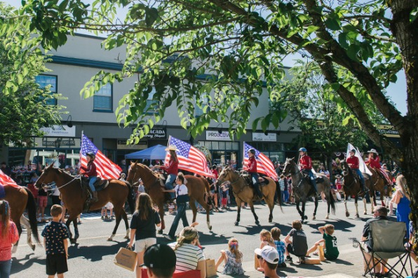A parade featuring riders on horseback carrying American flags, moving along a street lined with spectators.