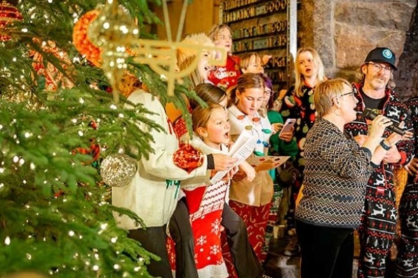 A group of people dressed in festive holiday sweaters, singing or reading near a decorated Christmas tree.