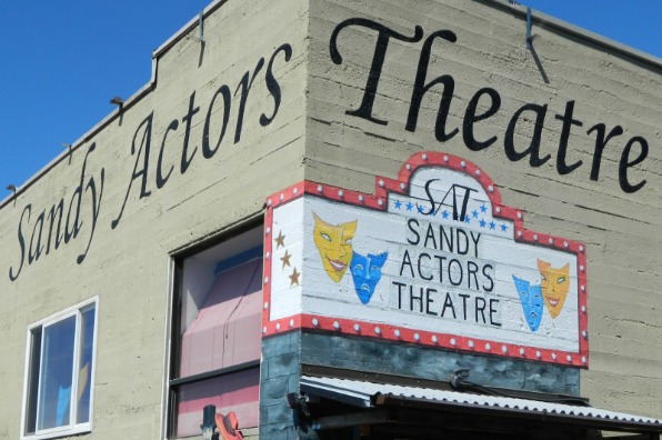 A building corner featuring signage for Sandy Actors Theatre, with the name painted on both walls and a marquee sign displaying comedy and tragedy masks in yellow and blue.