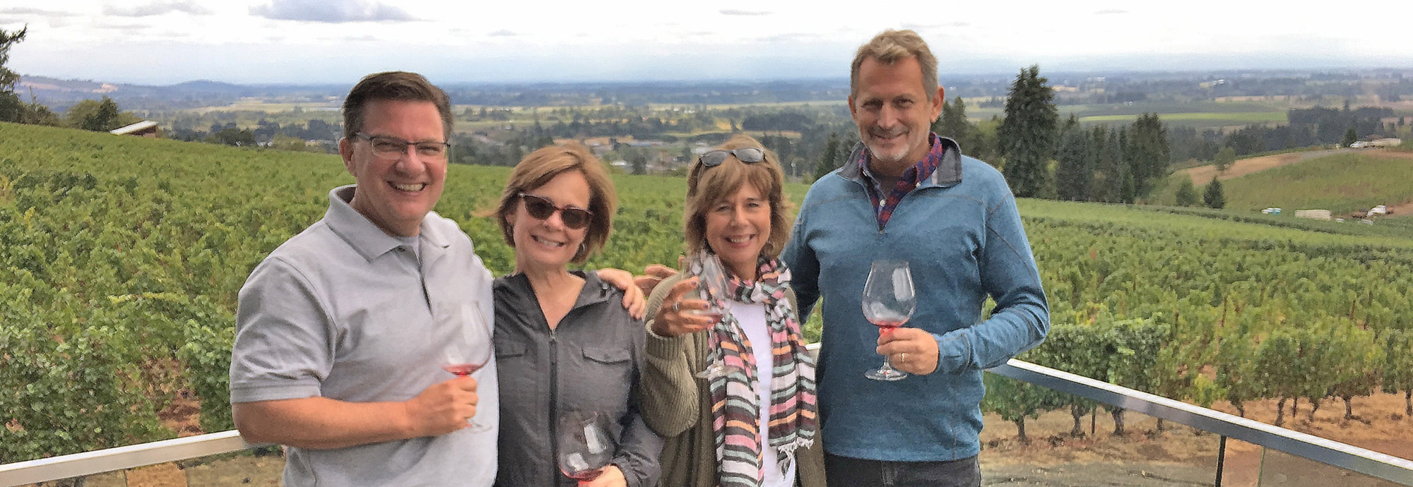 four people each holding a wine glass smile at camera with vineyard behind them