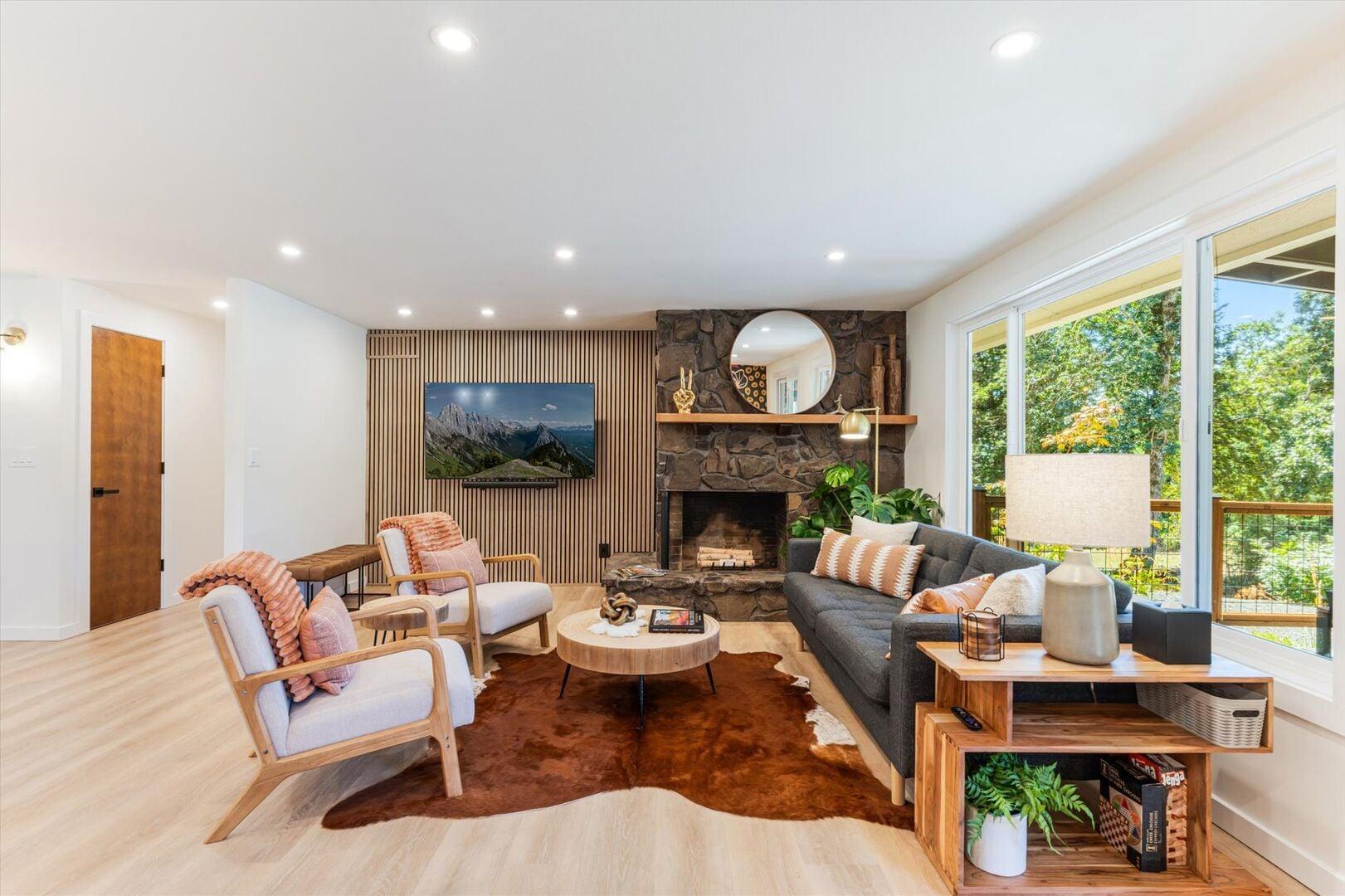 Modern living room with stone fireplace, flat-screen TV, and large windows with greenery outside.